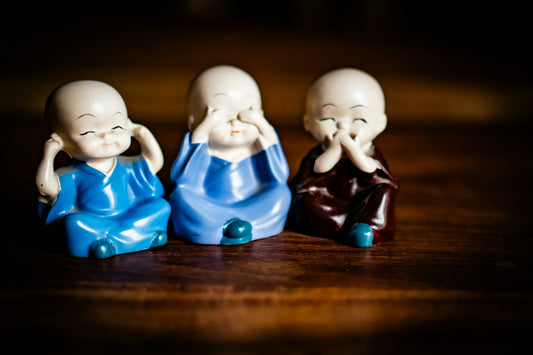 Three small monk figurines sitting on a wooden surface, each covering ears, eyes, or mouth